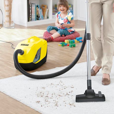 Keltainen pölynimuri ja imuroiva aikuinen valkoisella matolla, lapsi leikkii taustalla.