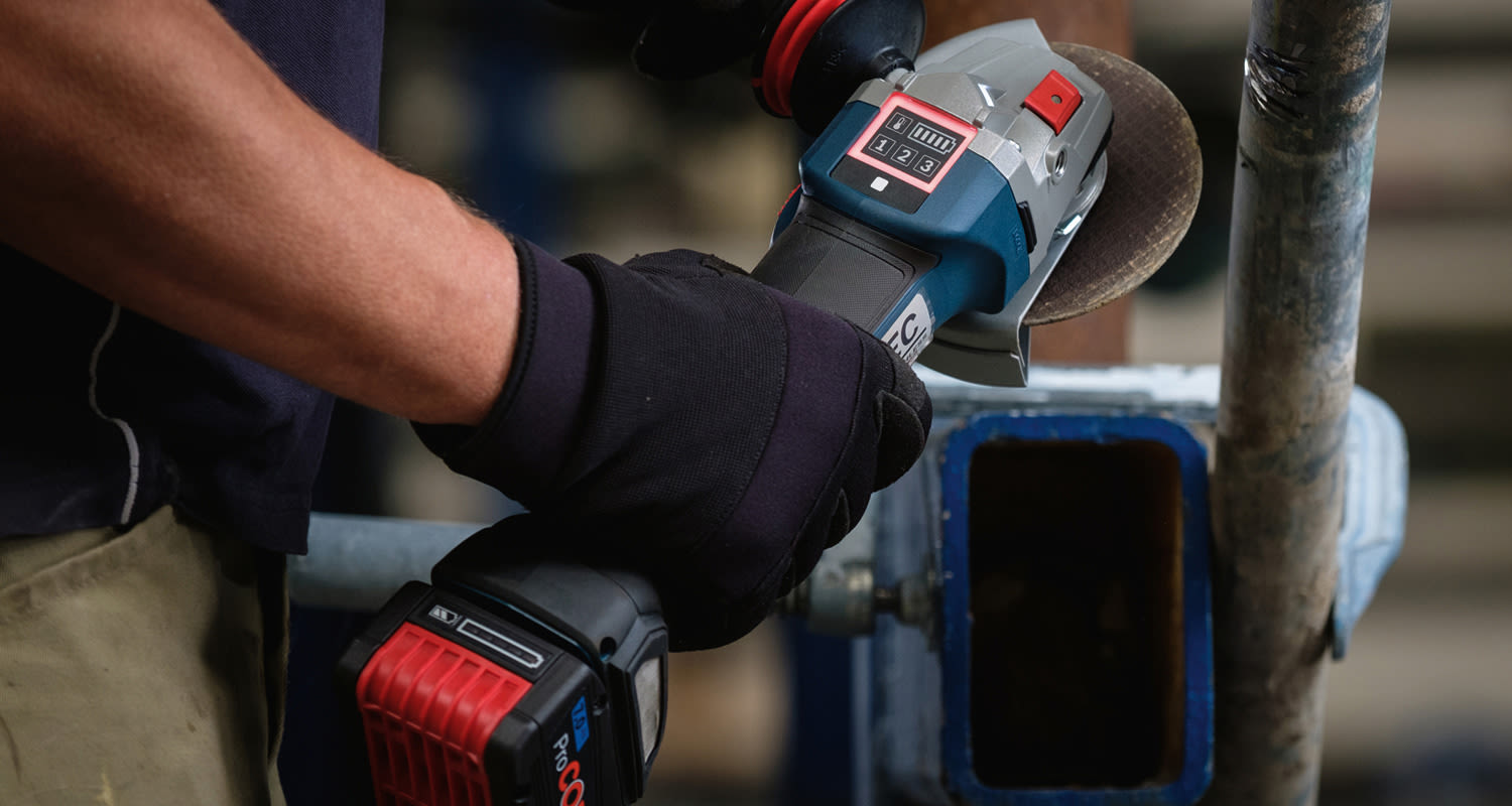 Mustakäsineinen käsi pitelee siniharmaata ja punamustaa akkuhiomakonetta metalliputken vieressä