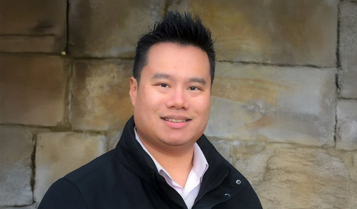 Smiling man with short black hair, wearing a black jacket and white shirt, posing in front of a stone background.