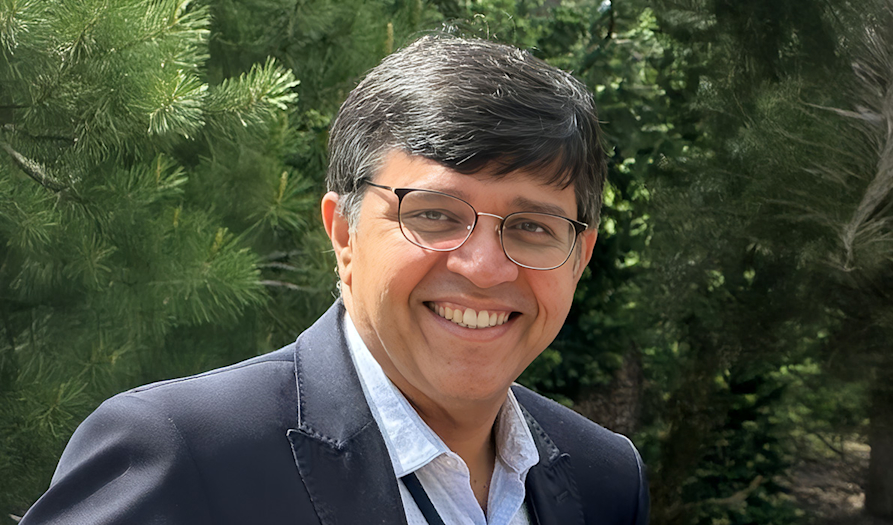 A smiling man with glasses and dark hair stands outdoors against a backdrop of lush green trees. He wears a dark jacket and light shirt, conveying a warm, friendly tone.