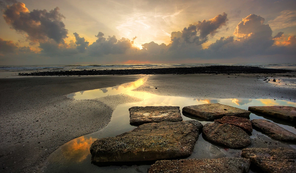 A serene beach scene at sunset, featuring rocks in the foreground and colorful clouds in the sky, reflecting on the water.