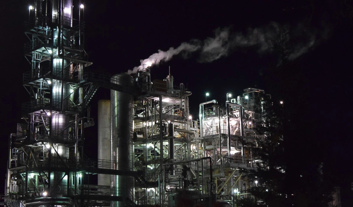 Industrial plant at night, featuring large metal structures emitting steam and illuminated by bright lights against a dark sky.