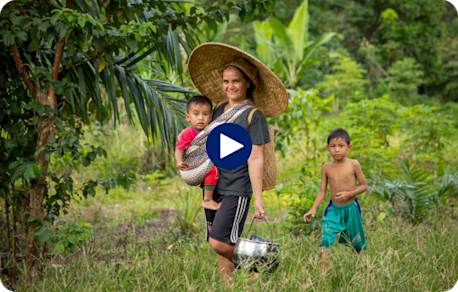 A woman is walking in a field carrying a child on her back, while another child walks nearby in a green area.