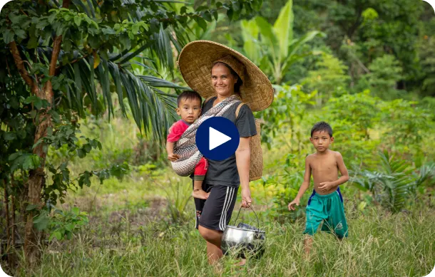 A woman is walking in a field carrying a child on her back, while another child walks nearby in a green area.