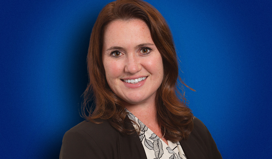 Smiling woman with long brown hair wearing a black blazer and patterned blouse against a blue background, showcasing a friendly demeanor