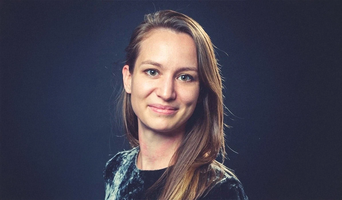 Portrait of a smiling woman with long brown hair, wearing a dark top, set against a dark background.