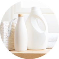 Bottles of laundry detergent and fabric softener on a wooden surface with towels and a basket in the background.