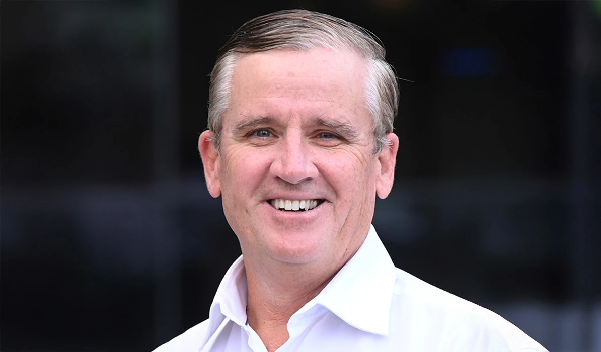 Smiling man with short hair, wearing a white collared shirt, standing in front of a blurred background.