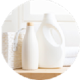 Bottles of laundry detergent and fabric softener on a wooden surface with towels and a basket in the background.
