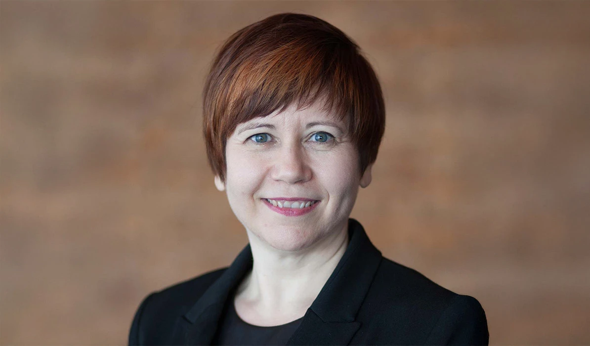 Smiling woman with short red hair, wearing a black blazer, posing in front of a neutral brown background.