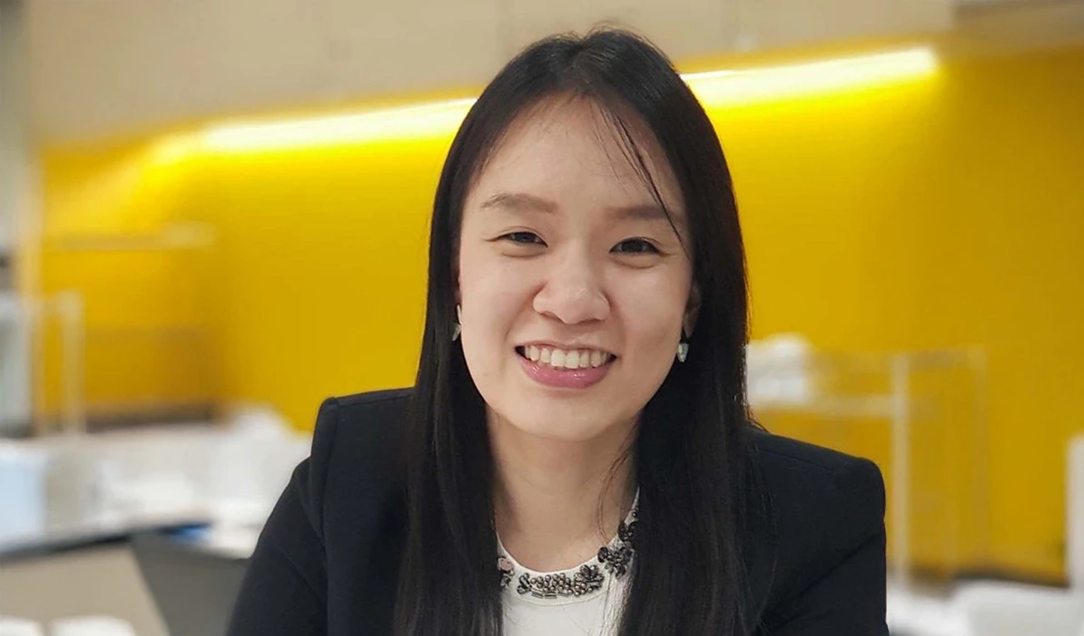 Smiling woman with long black hair, wearing a black blazer and a statement necklace, with a bright yellow background.