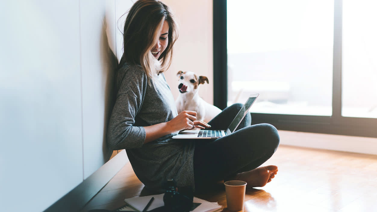 A woman sits with her legs crossed against a wall, using her laptop, accompanied by a dog.