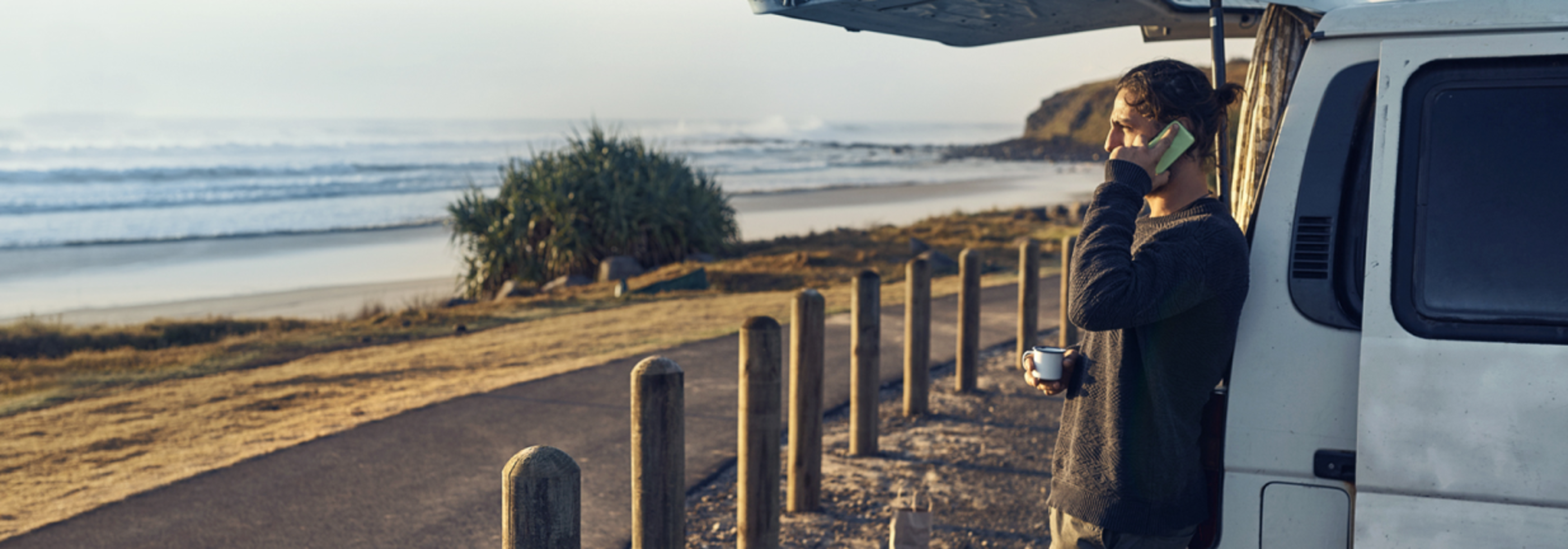 Man leaning on a van using a mobile phone by the beach