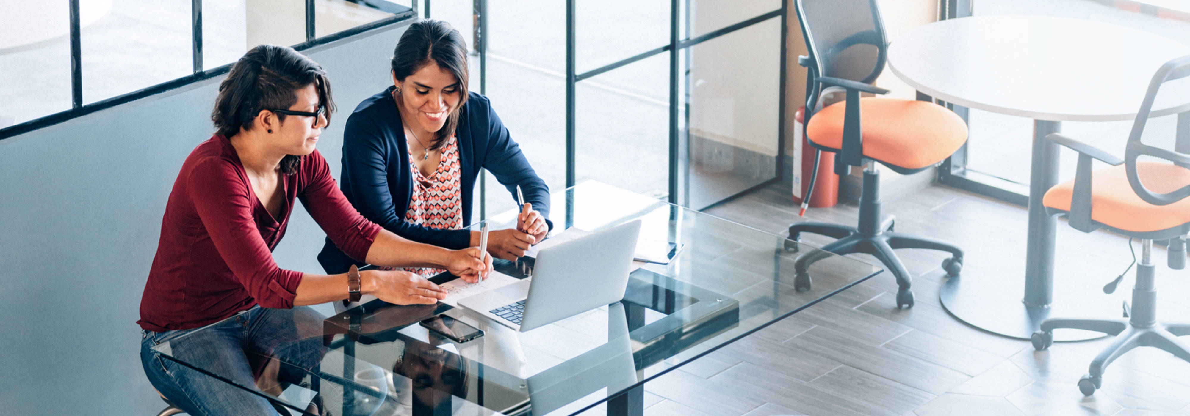 Two women at an office desk using a notepad and laptop