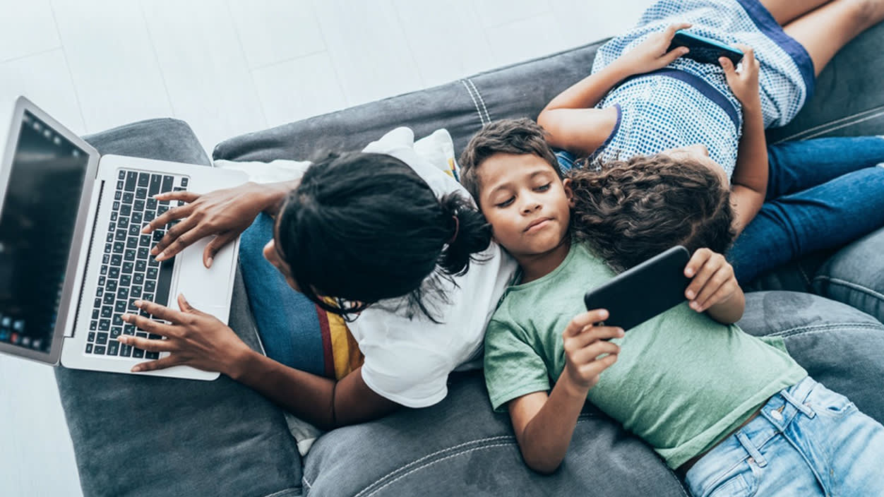 A mother and two kids lay on a couch together, each looking at their own device.