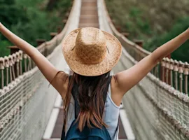 Ragazza su ponte tibetano. E' di spalle con le braccia alzate, indossa un cappello di paglia e uno zainetto. Il ponte si trova su una foresta tropicale