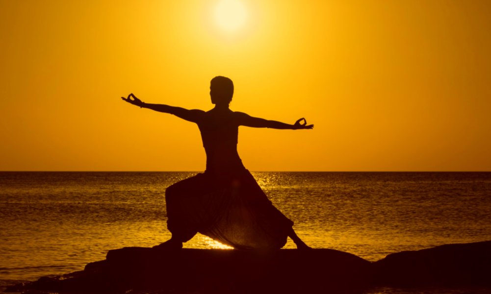 La silueta de una yogi posando al lado del mar con el cielo y sol amarillos atrás 