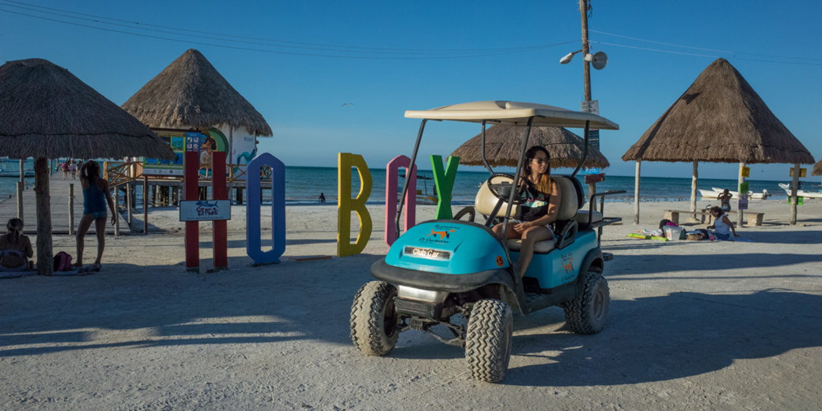 A young woman driving a rental golf cart next to the Isla Holbox wooden pier on a sunny day.