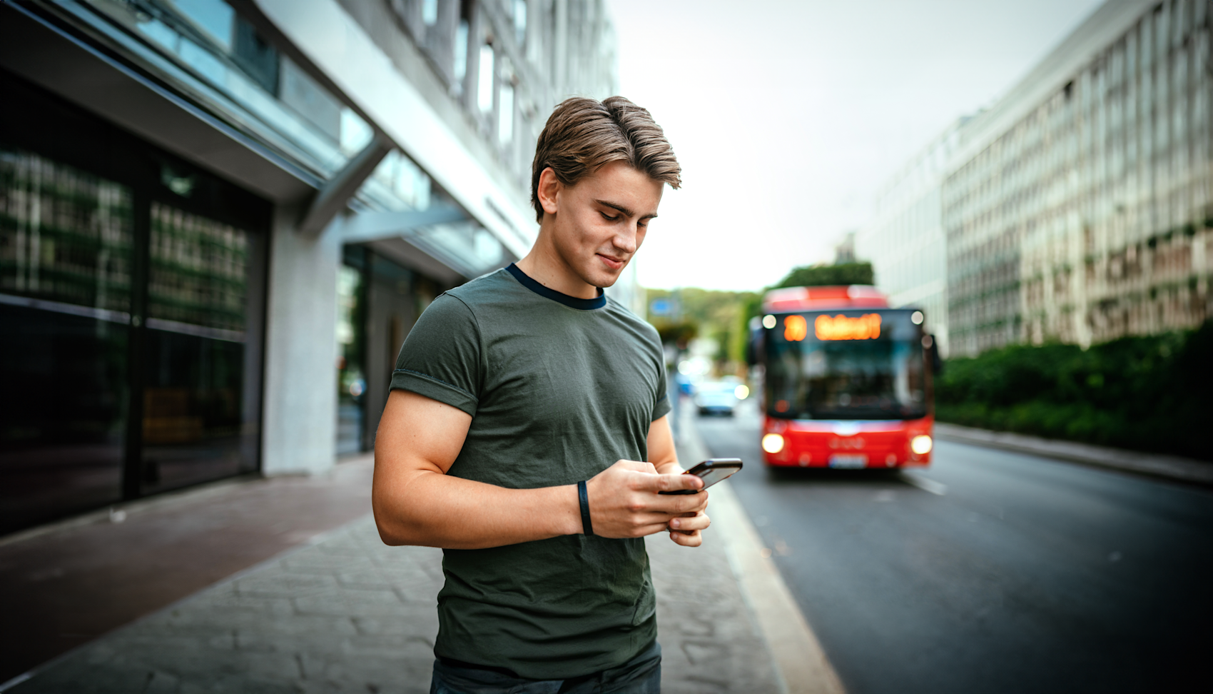 En ung mann som står på en fortau og ser på telefonen sin. I bakgrunnen er det en rød buss som kjører langs gaten. Han har på seg en grønn T-skjorte og virker å være fokusert på det han leser eller skriver på telefonen sin. Området rundt ser ut til å være en bygate med bygninger på hver side.