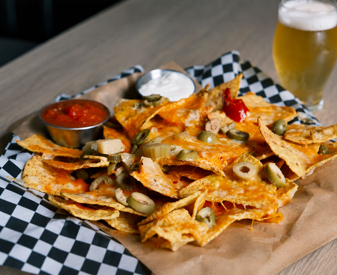 Une généreuse assiette de nachos gratinés garnis d’olives, accompagnée de salsa, de crème sure et d’une bière.