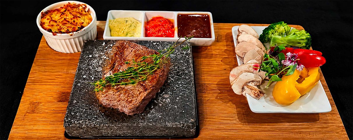 Steak sur pierre chaude avec herbes, gratin, sauces variées, légumes frais et champignons sur planche en bois.