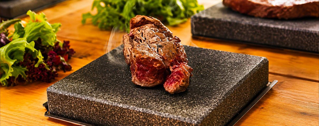Un steak saignant sur une pierre chaude, entouré de laitue verte et rouge, sur une table en bois.