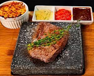 Steak sur pierre noire avec herbes, gratin doré à gauche, trois sauces colorées en arrière-plan sur planche en bois.