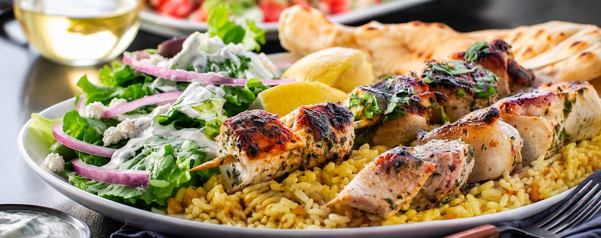 Une savoureuse brochette de poulet grillé, servie avec du riz parfumé, une salade grecque fraîche, du pain pita et une coupe de vin blanc.