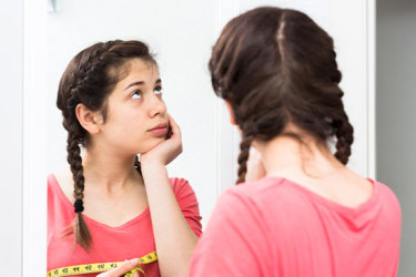 Uma adolescente a pensar em frente do espelho a medir o perímetro das suas mamas. Olha para cima suportando o queixo com a mão