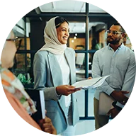 A smiling woman wearing a light-colored hijab holds a stack of papers, surrounded by an engaged group of people in an office environment. A blue circle outline surrounds the icon.