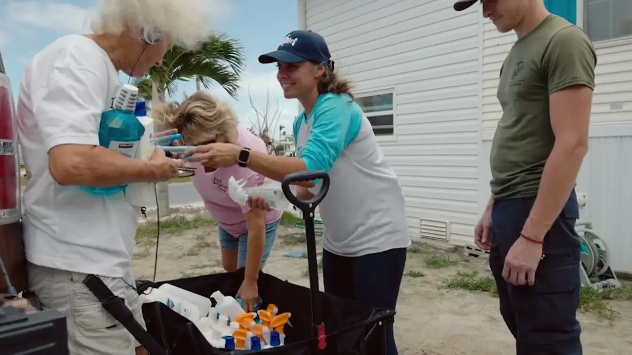 Volunteers distribute cleaning and hygiene products to community members outside a storm-damaged home.