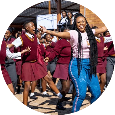 A Black woman with long, dark braided hair. She leads a larger group of female students in dance. The girls wear red and white uniforms.