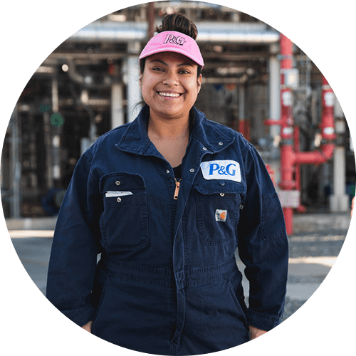 Woman smiling in a manufacturing facility, wearing a pink visor, dark blue jumpsuit with P&G logo.