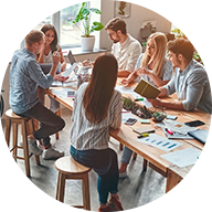 A group of six people engaged in a discussion sit around a wooden table with papers, notebooks, and electronic devices spread out. A blue circle outline surrounds the icon.