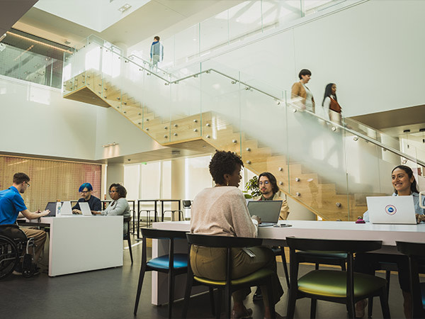 Several people sit together at various office work stations. Two women walk down a staircase in the background.