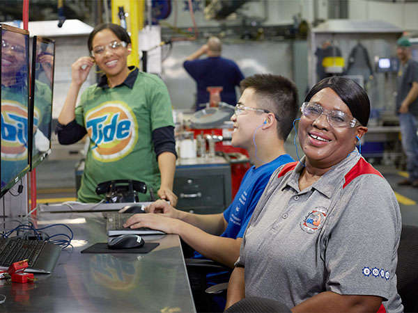 Two women and a man sit in front of several computers in a manufacturing plant. They wear protective eye gear and smile at the camera.