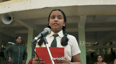 A young Indian girl in a white and green school uniform. She stands at a microphone while holding a red piece of paper.