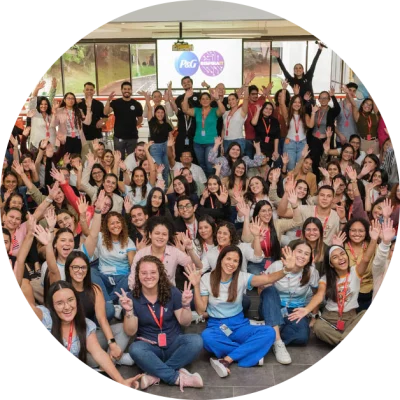 A large group of Hispanic Procter & Gamble employees pose, with many sitting on the floor and others standing in the background. They are waving and smiling. The group is predominantly female.