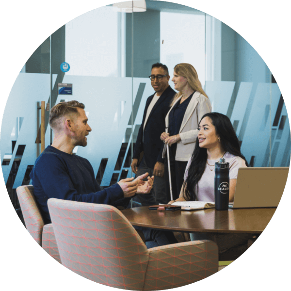 A man and woman are working together at a conference table, talking together. Another man and woman are walking outside the glass-walled room, and another man works at a separate table, his back to the others.