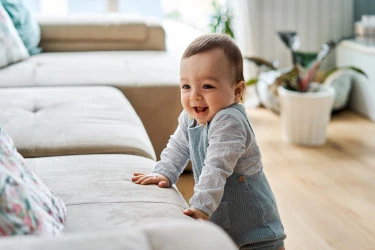 A smiling baby holds themself up by a couch cushion