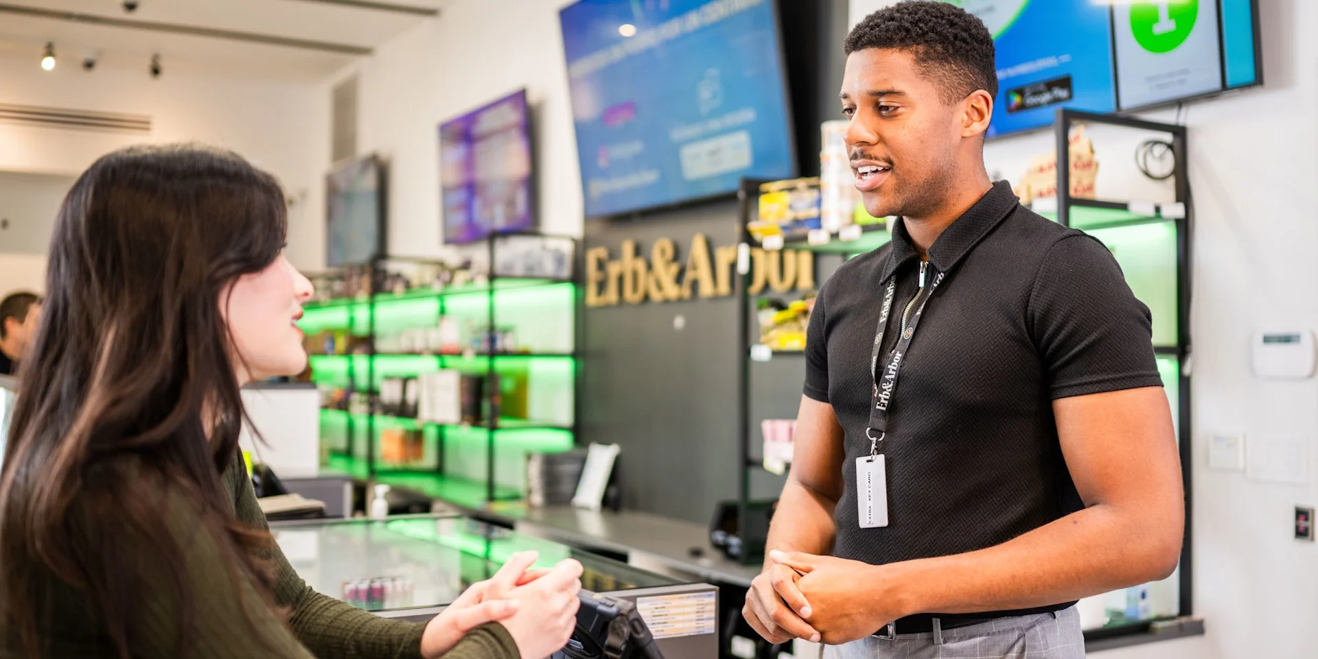 Dispensary budtender in a branded polo talking with a customer at the sales counter