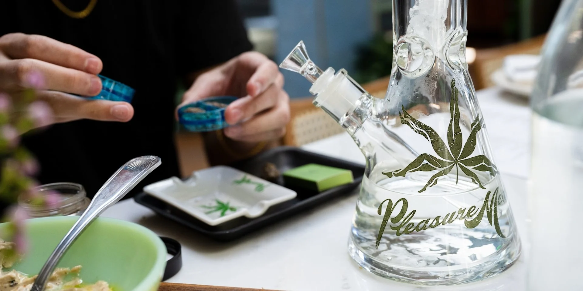 A customer grinding cannabis flower at a table set with a rolling tray, ashtray, and branded glass water pipe.