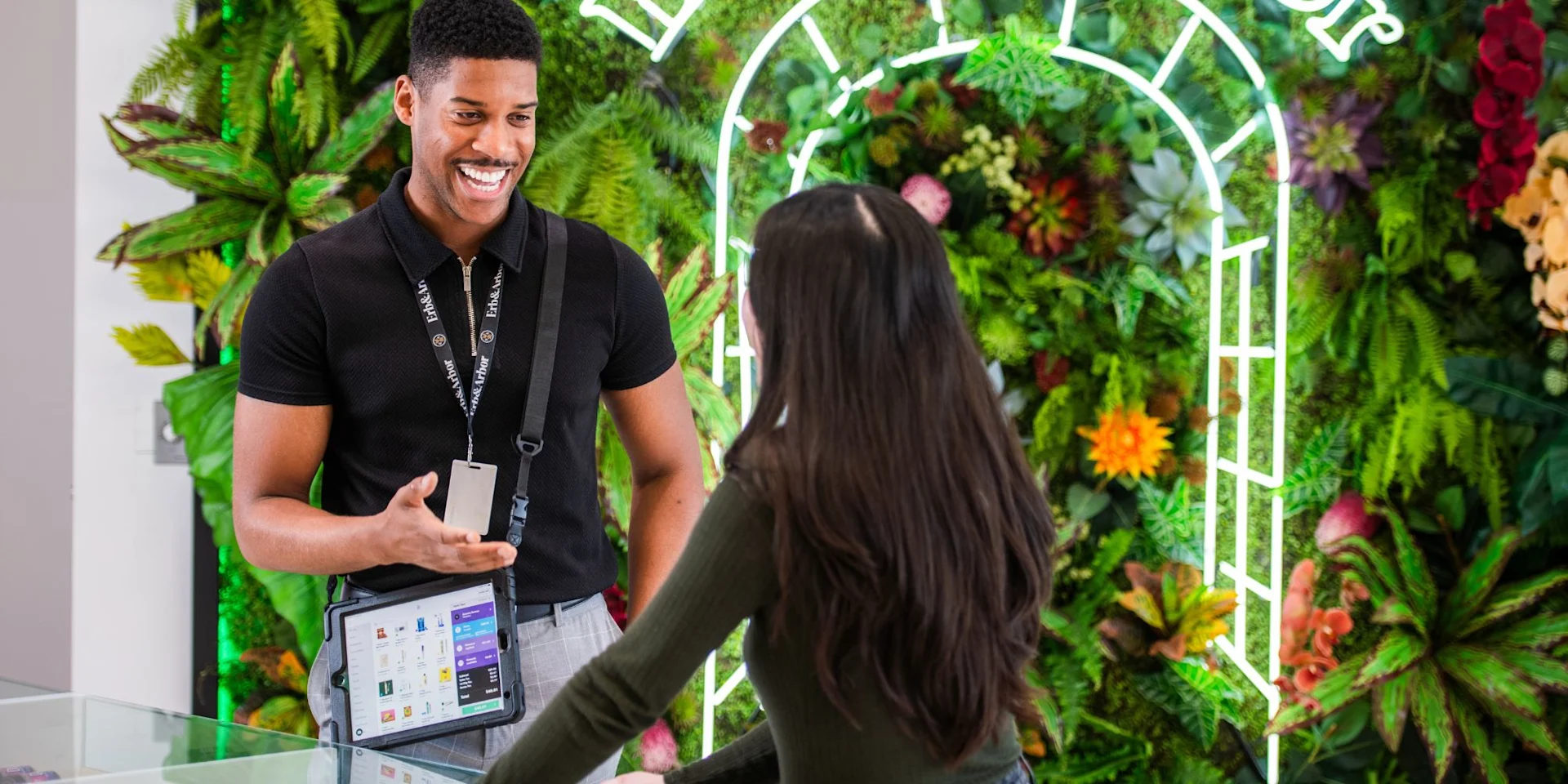 Smiling budtender using a tablet POS to check out a customer in front of a live plant wall