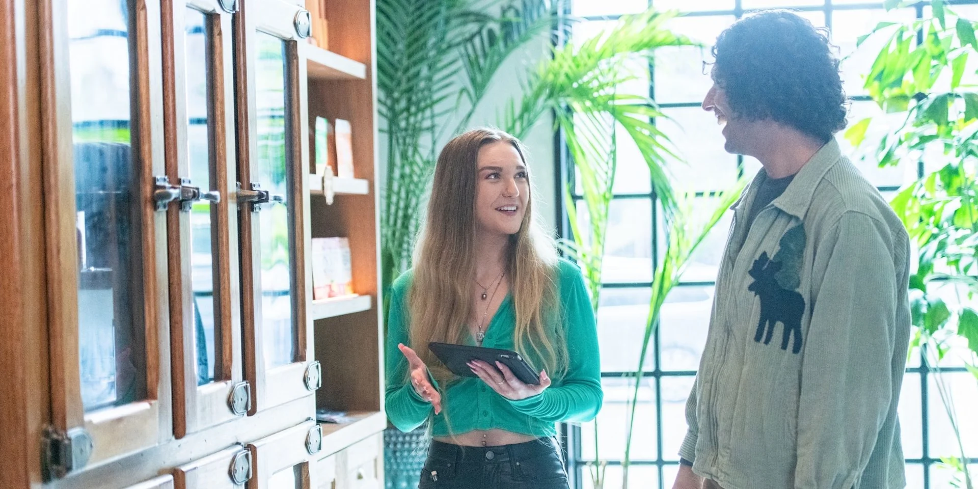 A dispensary staff member with a tablet talks with a customer beside product shelving in a bright, plant-filled retail space.