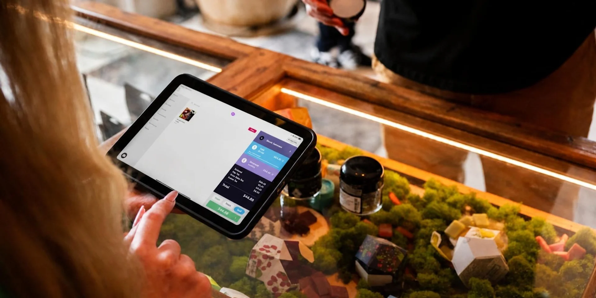 A budtender holds an iPad running Meadow's point of sale, reviewing an order summary and total at a lit dispensary display case.