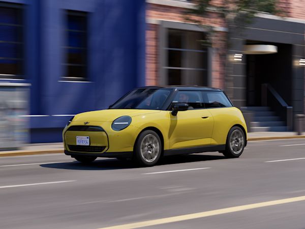 Yellow MINI Cooper electric with black accents driving on a city street, with blue and brick buildings and greenery in the background.