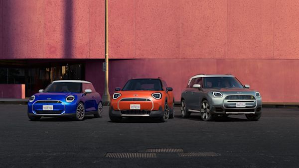 Three MINI cars parked side by side in front of a pink wall. From left to right, the cars are blue, orange, and silver.