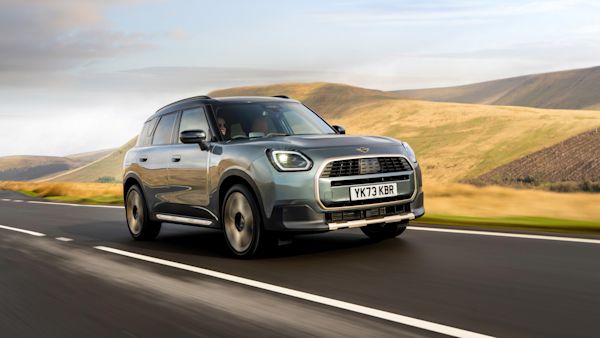 A silver MINI Countryman Electric driving on a scenic road with rolling hills and a partly cloudy sky in the background.