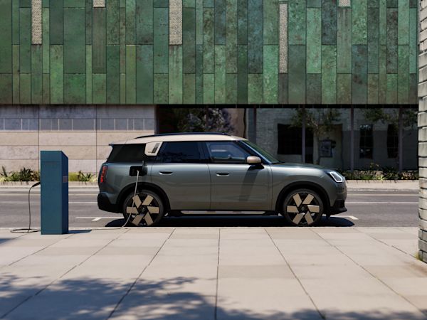 A grey MINI Countryman electric parked and charging at a public charging station on a city street, with a modern green-tiled building in the background.