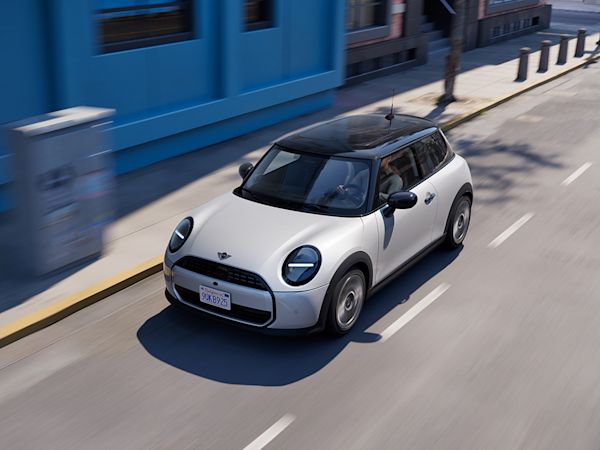 A white MINI Cooper with a black roof driving on a city street, with a blue building and large windows in the background.